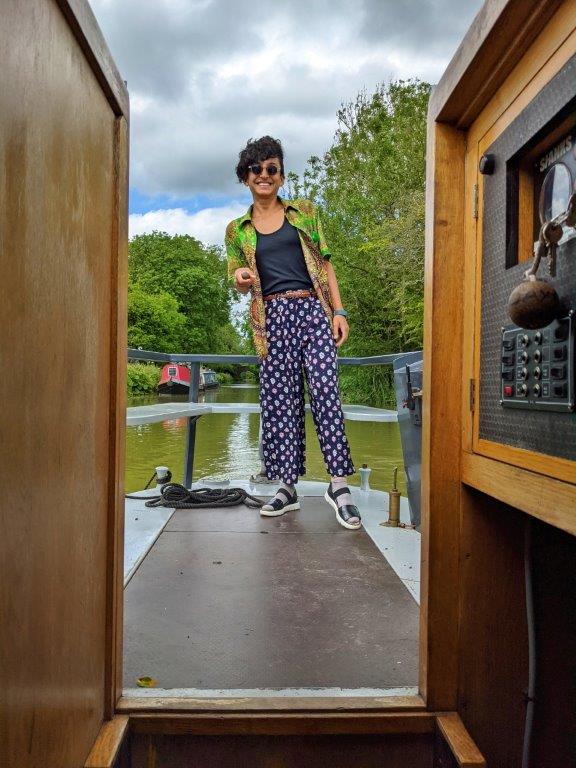 A woman in green shirt and sunglasses and a big smile steering the tiller of a narrowboat