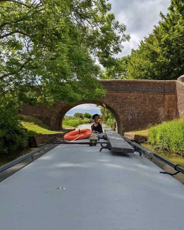 A view down the length of the roof of a narrowboat passing under the arch of a bridge, with a woman at the far end steering and waving to the camera