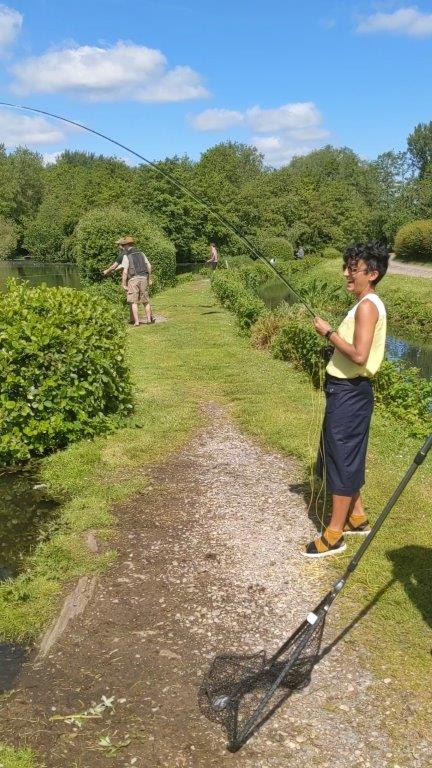 A woman in a sleeveless white and yellow top, sunglasses and a long navy skirt fishing with a rod by the side of a lake