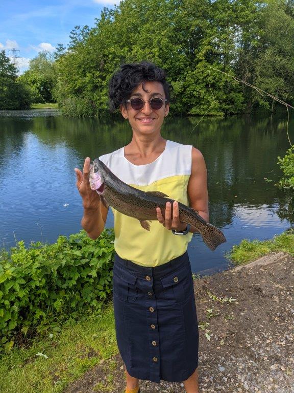 A woman in a sleeveless white and yellow top, sunglasses and a long navy skirt proudly holding up a trout she caught