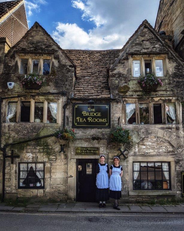 A medieval looking three storey stone buildings with 'The Bridge Tea Rooms' above the entrance, two young women in tea maid outfits standing out the front