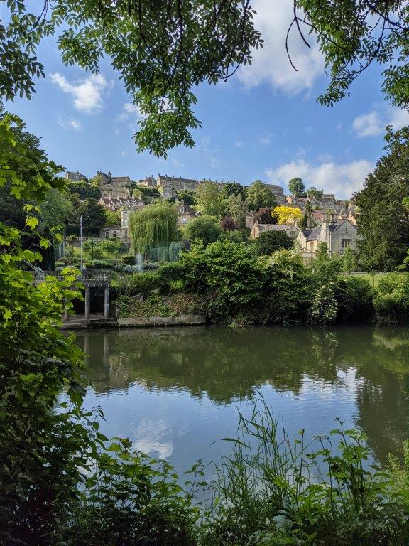 View of a still river with lush vegetation and beautiful stone buildings on the opposite bank, image framed by tree branches and foliage in the foreground