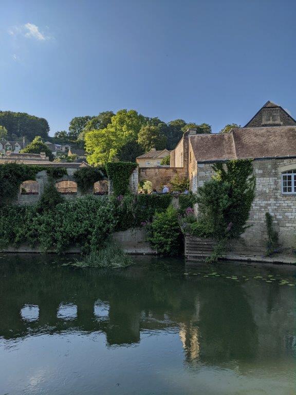 View across the width of a peaceful river, old stone buildings and lots of climbing vegetation on the opposite bank