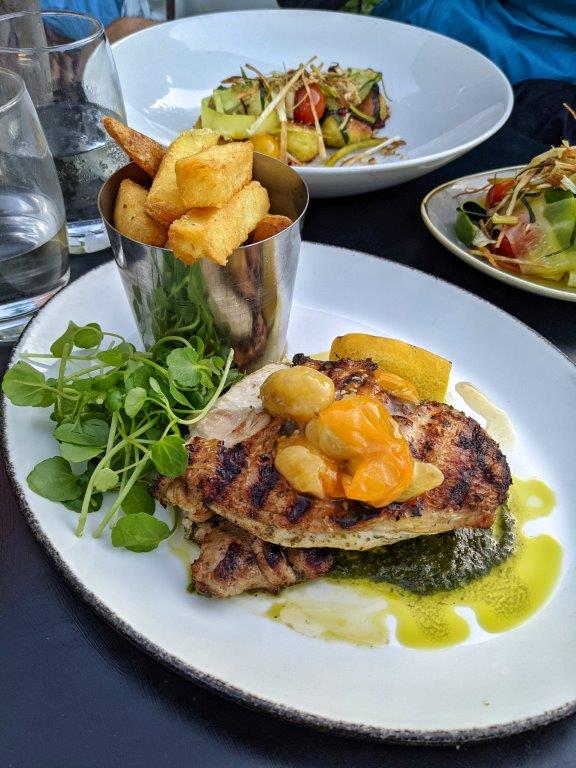 A plate of charred chicken with confit yellow tomatoes on a green sauce with a pile of chunky chips