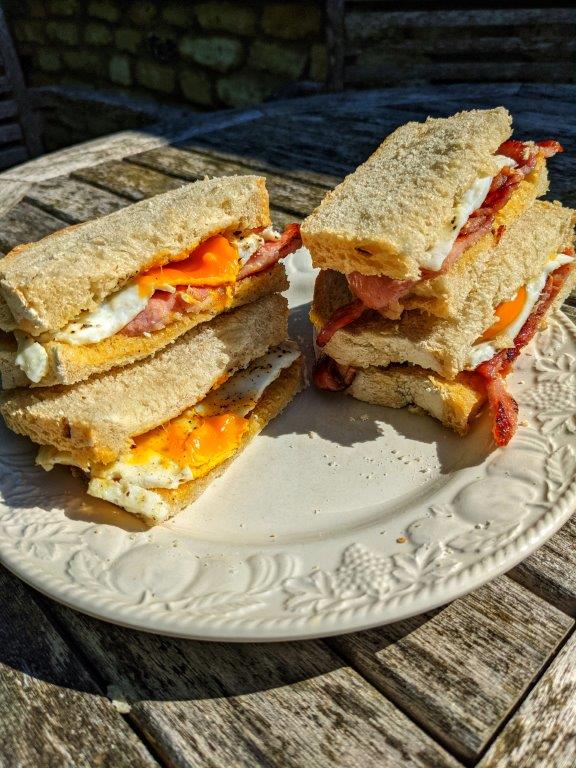 Two bacon and egg sandwiches cut in half and stacked on white bread, on a white plate outside in the sunshine
