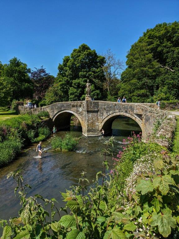 A river on a sunny day with children splashing in the shallows, an old stone bridge with two arches passing over it