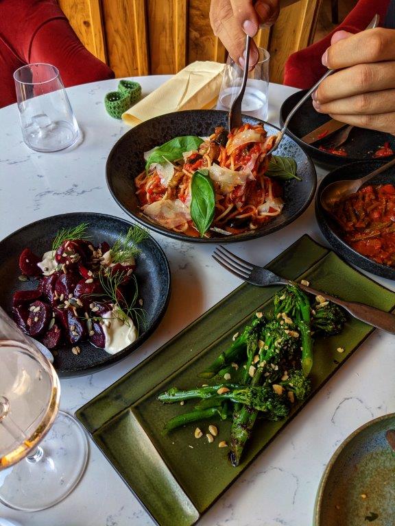 An assortment of dishes laid out on a white marble table with two hands forking up a big bowl of pasta in the middle