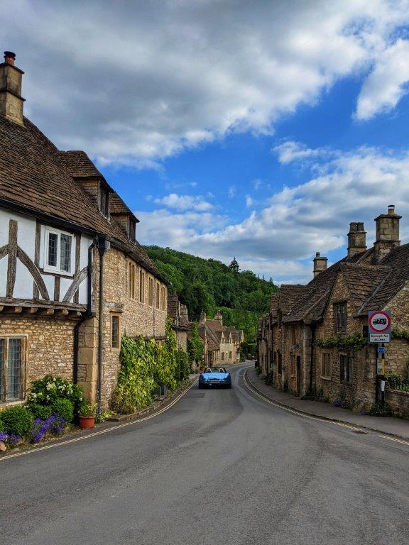 View down an English village road lined with beautiful old houses, with an electric blue sports car zooming off into the distance