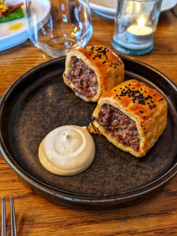 Two halves of a well filled artisan sausage roll next to a dollop of sauce arranged on a black plate on a wooden table
