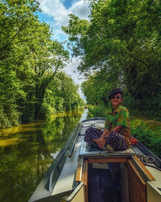 A girl in a green short sitting crossed legged on the roof of a narrowboat moored on a peaceful canal surrounded by lush green trees