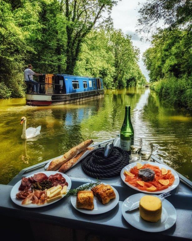 A delicious array of picnic hamper edibles arranged at the front of a narrow boat against a backdrop of a peaceful canal with a swan and passing narrowboat